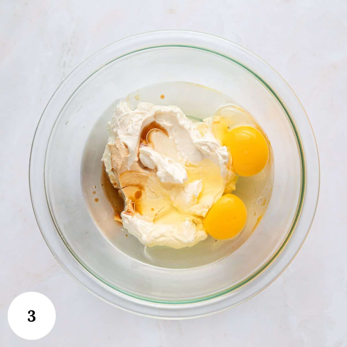 A glass mixing bowl containing ingredients for blueberry yogurt muffins-two raw eggs, cream cheese, and vanilla extract-rests on a light countertop. A white circle with the number 3 appears in the bottom left corner.