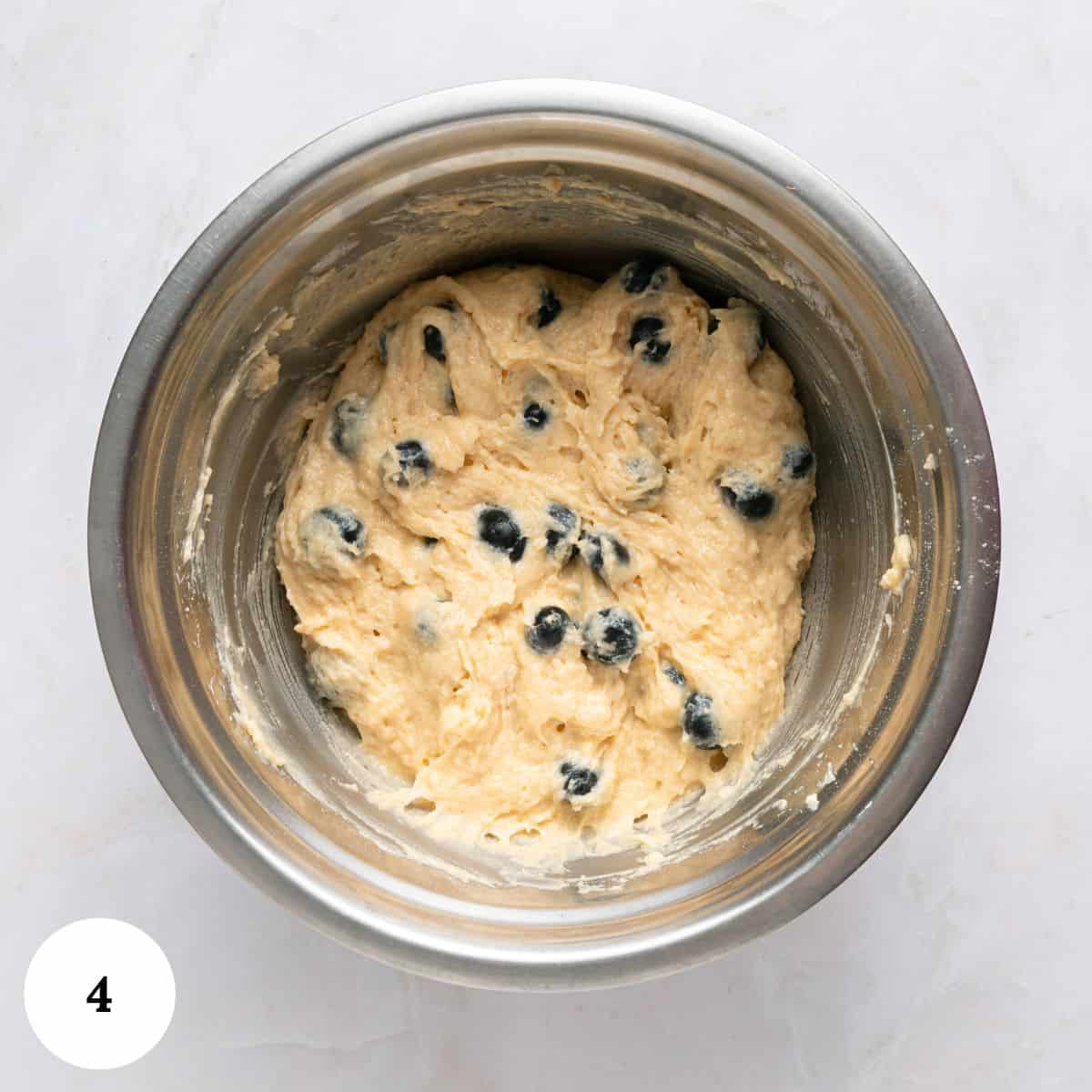 A stainless steel mixing bowl holds a thick, creamy batter dotted with blueberries for blueberry yogurt muffins. The bowl sits on a light marble surface, and a white circle with the number 4 appears in the lower left corner.