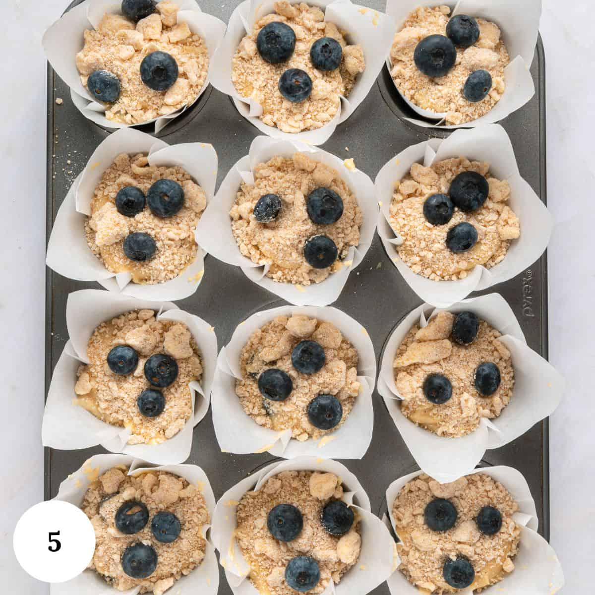 A muffin tray lined with white parchment paper holds twelve unbaked blueberry yogurt muffins topped with streusel and fresh blueberries. A white circle with the number 5 is in the lower left corner.
