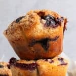 A close-up of golden-brown blueberry yogurt muffins with a crumbly topping and visible blueberry filling, stacked on top of each other against a light background.