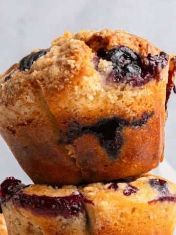 A close-up of golden-brown blueberry yogurt muffins with a crumbly topping and visible blueberry filling, stacked on top of each other against a light background.