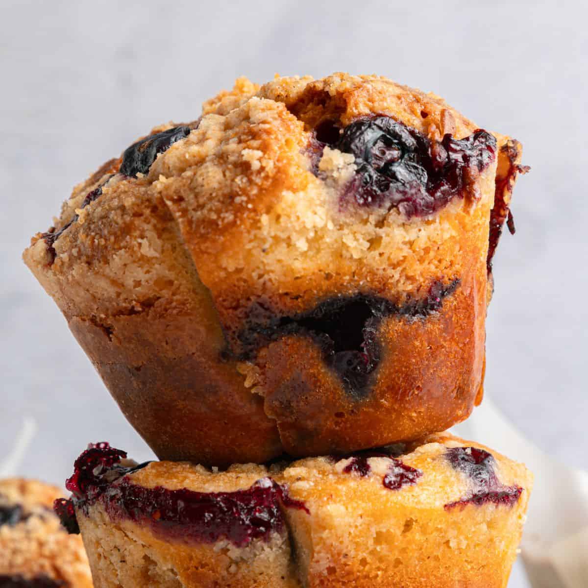 A close-up of golden-brown blueberry yogurt muffins with a crumbly topping and visible blueberry filling, stacked on top of each other against a light background.