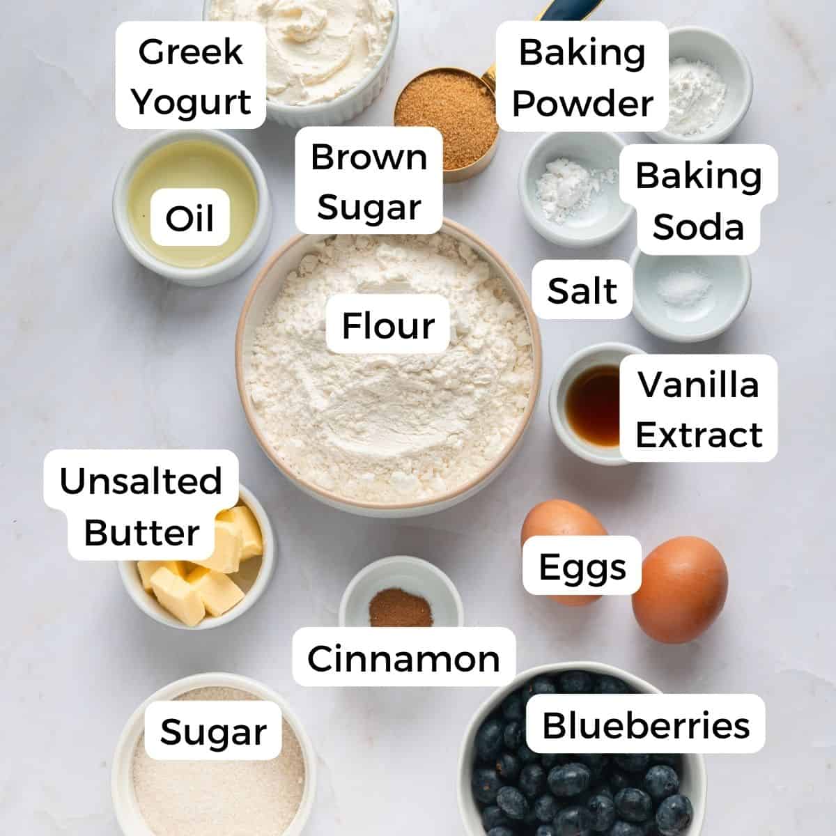 Various labeled bowls containing ingredients for baking blueberry yogurt muffins, including flour, brown sugar, Greek yogurt, eggs, blueberries, baking powder, baking soda, salt, cinnamon, vanilla extract, unsalted butter, oil, and sugar.