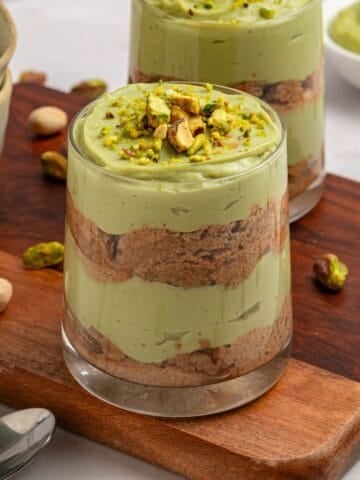 A glass filled with layers of creamy green pistachio tiramisu mousse and brown biscuit, topped with chopped pistachios, sits on a wooden board surrounded by whole pistachios and another dessert in the background.