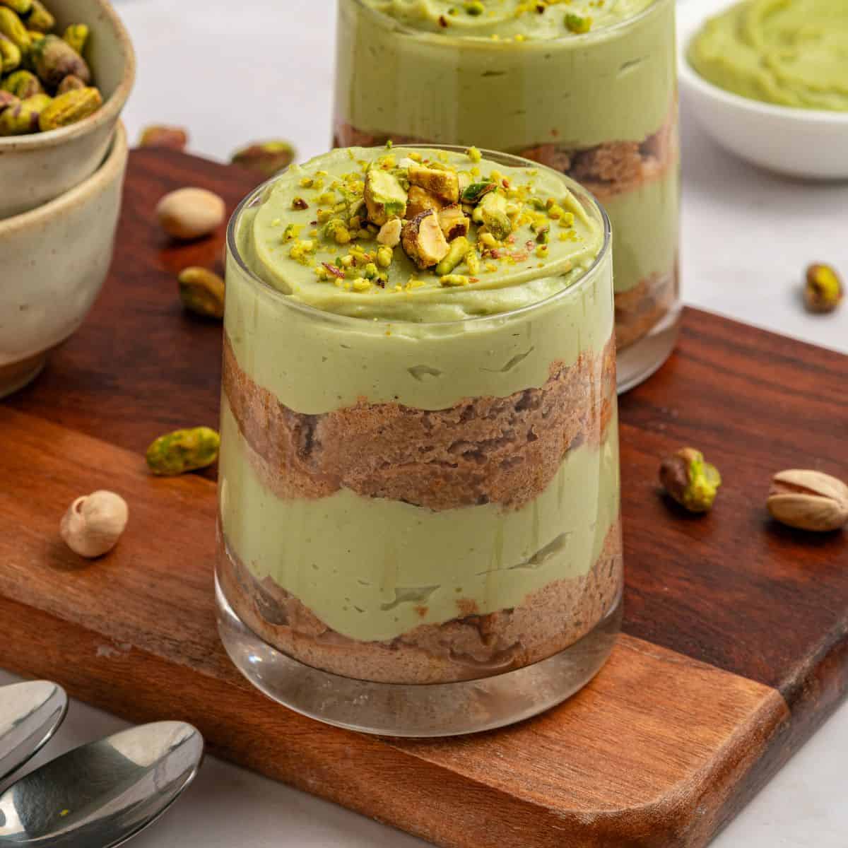 A glass filled with layers of creamy green pistachio tiramisu mousse and brown biscuit, topped with chopped pistachios, sits on a wooden board surrounded by whole pistachios and another dessert in the background.