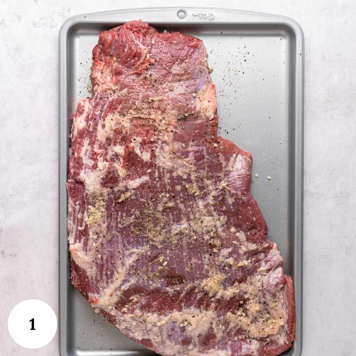 A raw oven roasted brisket seasoned with minced garlic, salt, and pepper sits on a metal baking sheet. The brisket is uncooked and spread out, with visible marbling and seasoning. A white circle labeled 1 is in the bottom left corner.