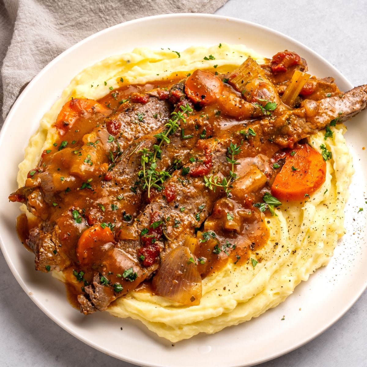 A plate of mashed potatoes topped with oven roasted brisket beef stew containing carrots, onions, celery, tomatoes, and fresh herbs, all covered in rich brown gravy.