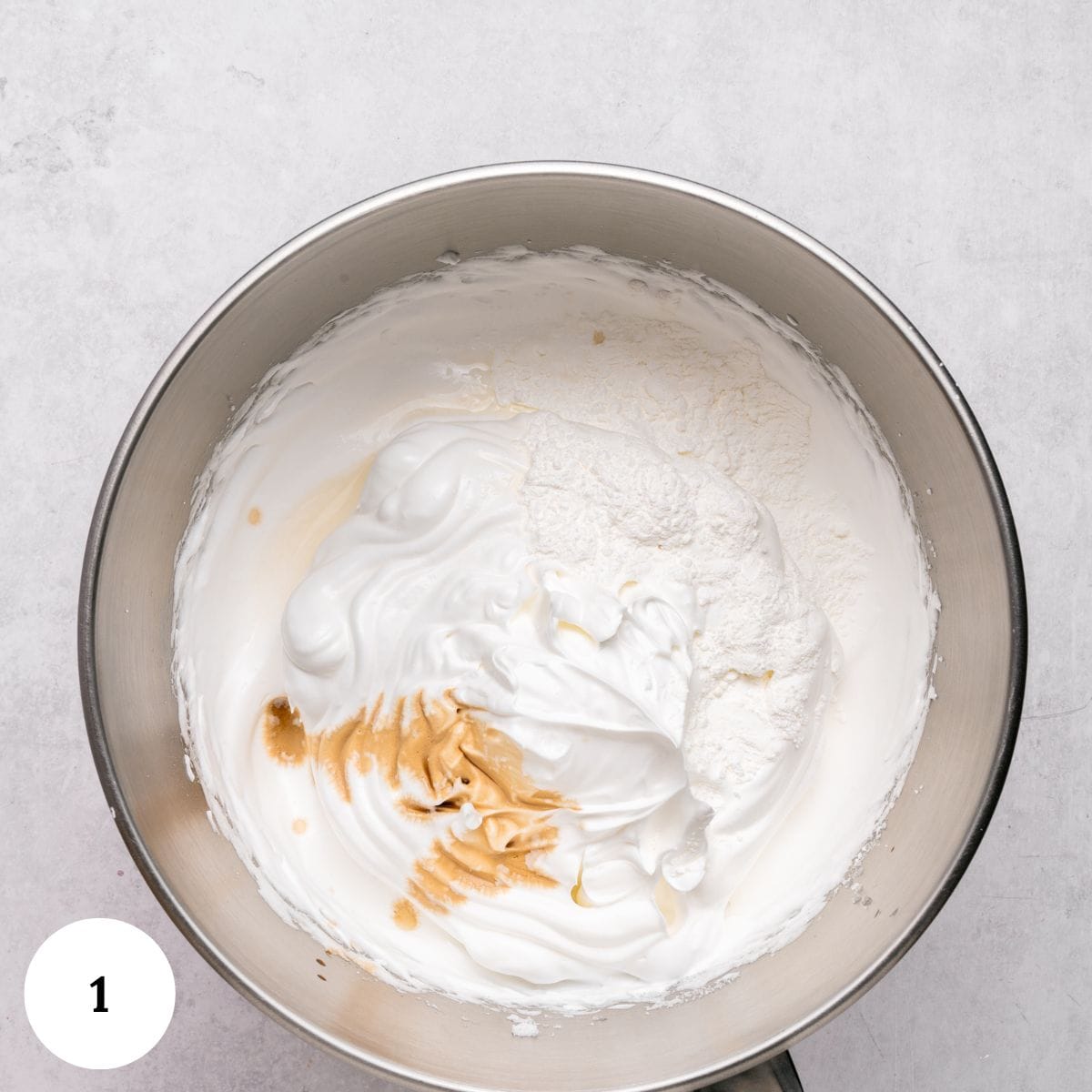A metal mixing bowl filled with fluffy whipped egg whites, flour, and a splash of vanilla extract for berry pavlova, viewed from above on a light gray surface. A white circle with the number 1 is in the lower left corner.