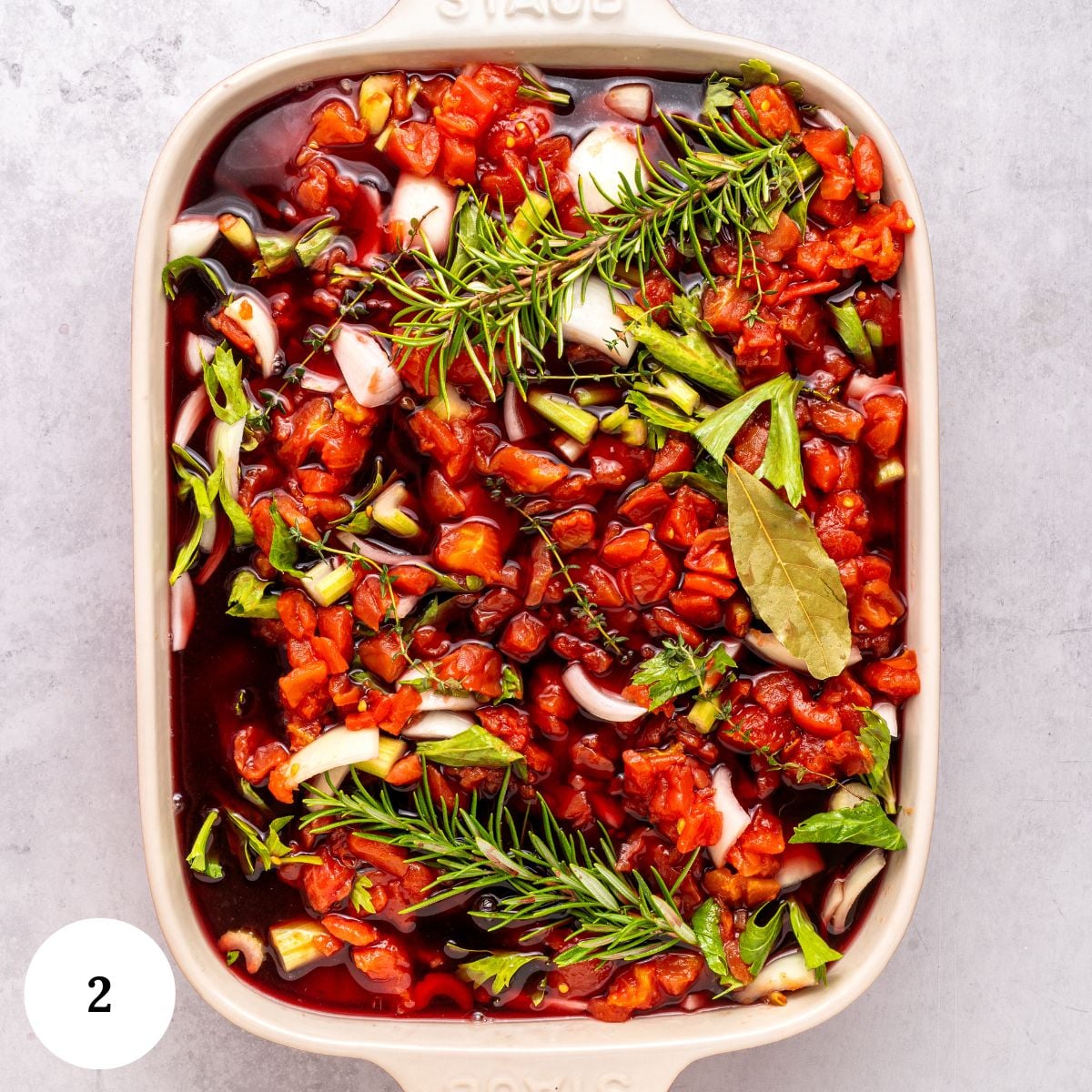 A baking dish filled with chopped tomatoes, onions, green herbs, and bay leaves in a dark red liquid sits ready for oven roasted brisket, on a light gray surface.