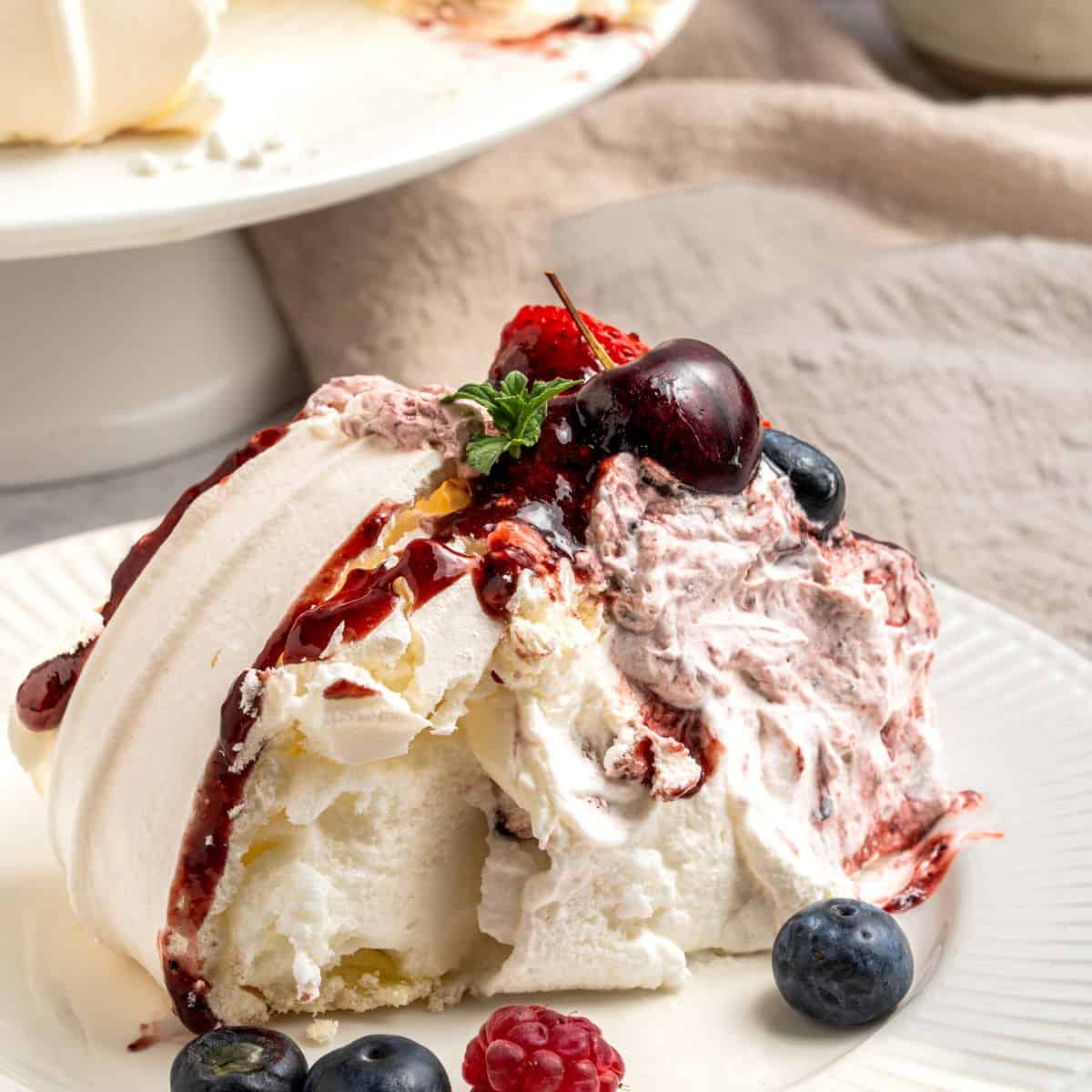 A slice of berry pavlova topped with whipped cream, berry sauce, and fresh berries including blueberries, raspberry, and a cherry, served on a white plate with extra berries on the side.