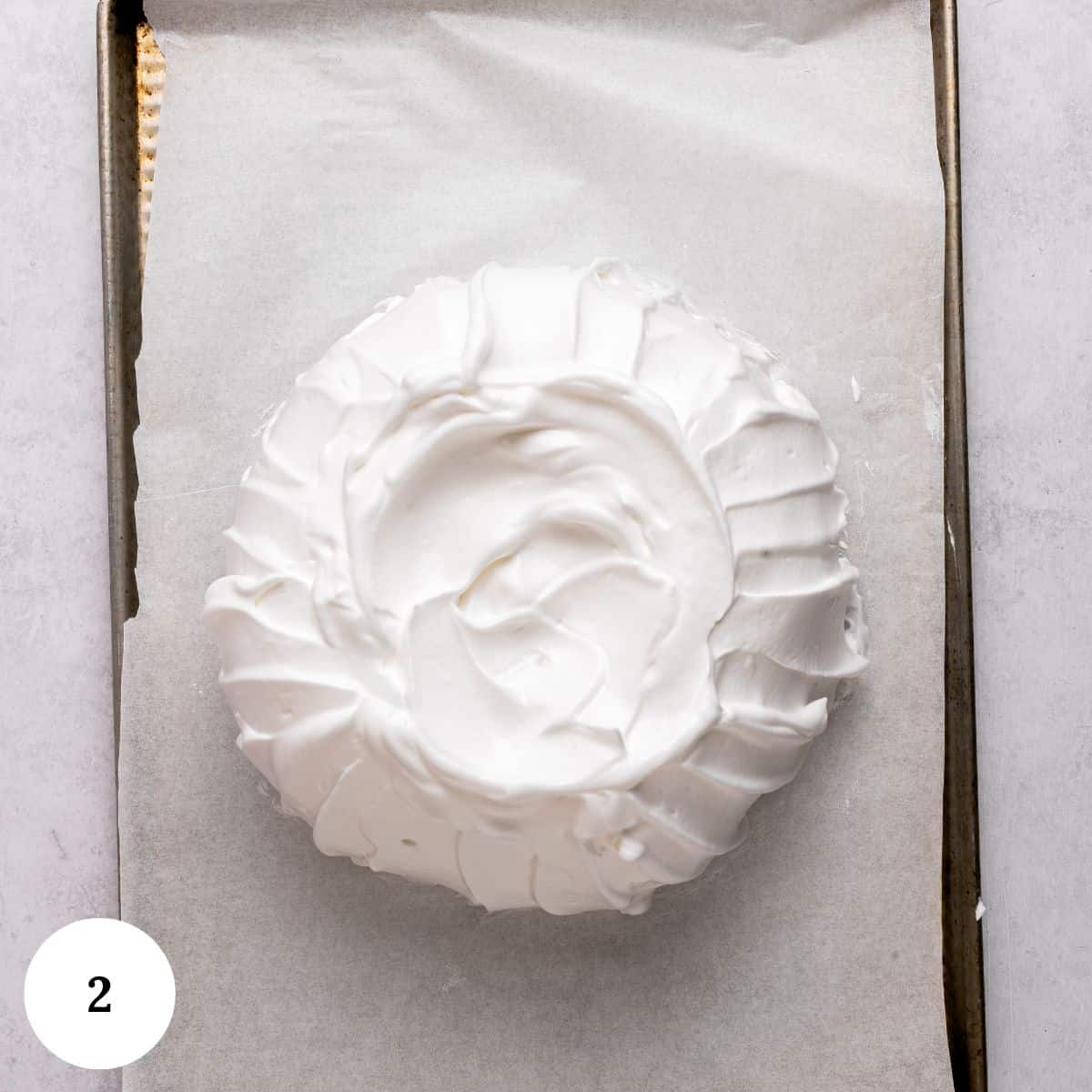 A round mound of fluffy white meringue with swirled edges sits on parchment paper on a baking sheet, ready to become a gorgeous berry pavlova. A small white circle with the number 2 is in the bottom left corner.