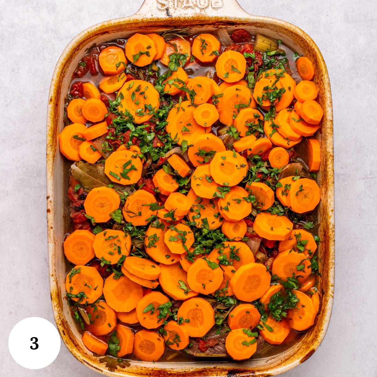 A rectangular baking dish filled with sliced carrots, herbs, and other vegetables in a cooked mixture-perfect as a side for oven roasted brisket-garnished with chopped parsley. A white circle with the number 3 is in the bottom left corner.