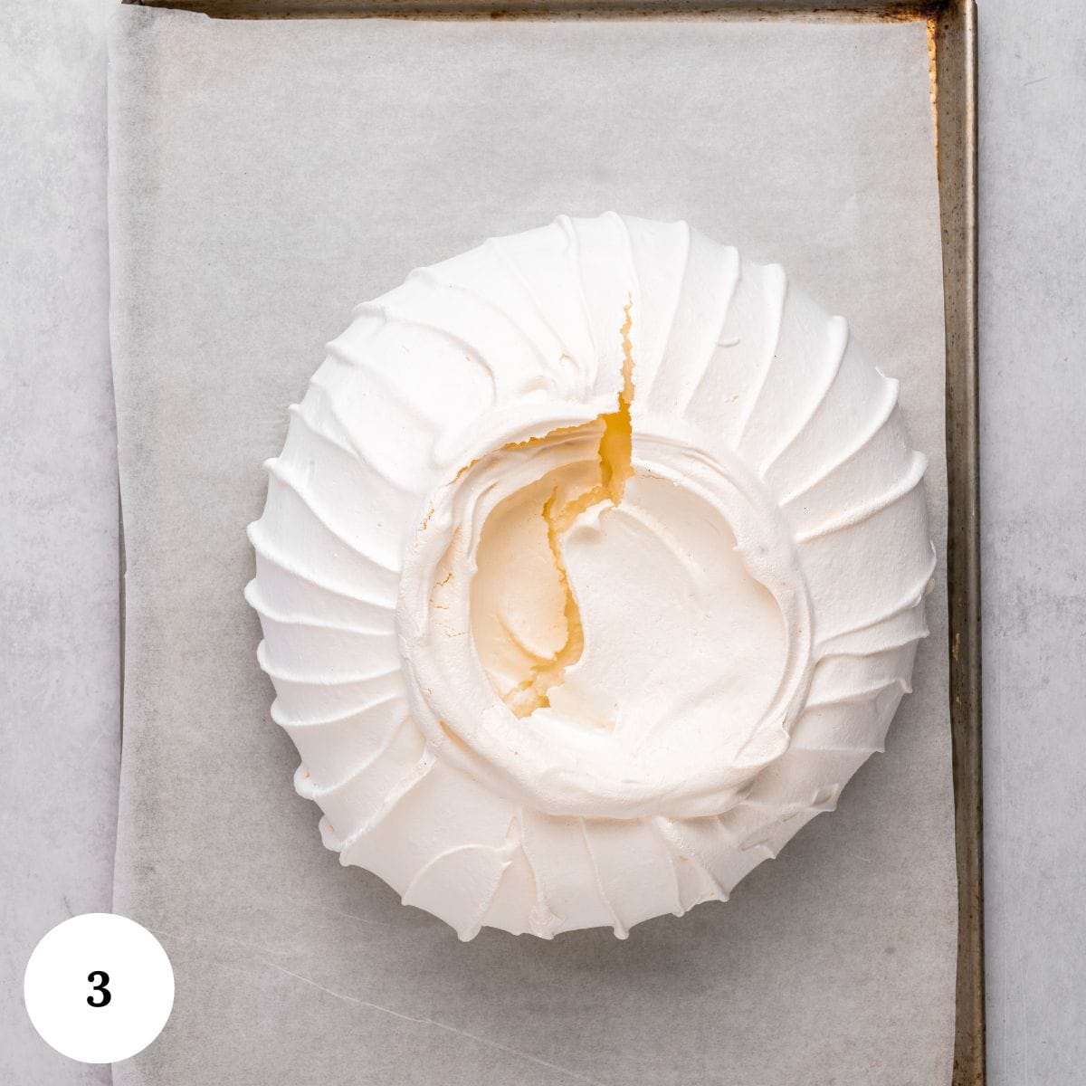 A round, white berry pavlova with a cracked center sits on parchment paper atop a baking sheet. A 3 in a white circle appears in the bottom left corner.