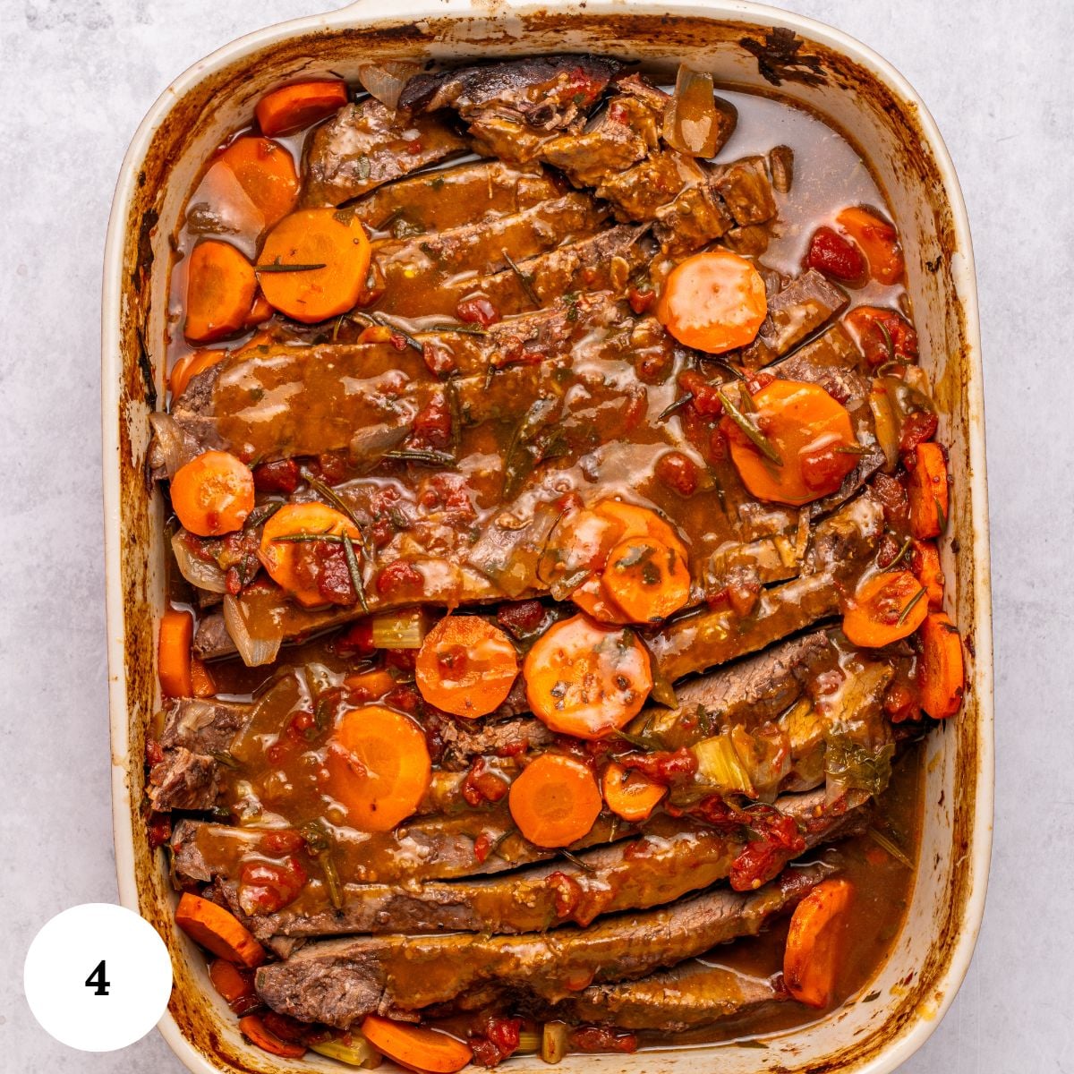 Oven roasted brisket is sliced in a casserole dish, topped with tomato-based sauce, carrots, celery, and herbs. The tender meat sits in rich gravy. A white circle with the number 4 is in the lower left corner.
