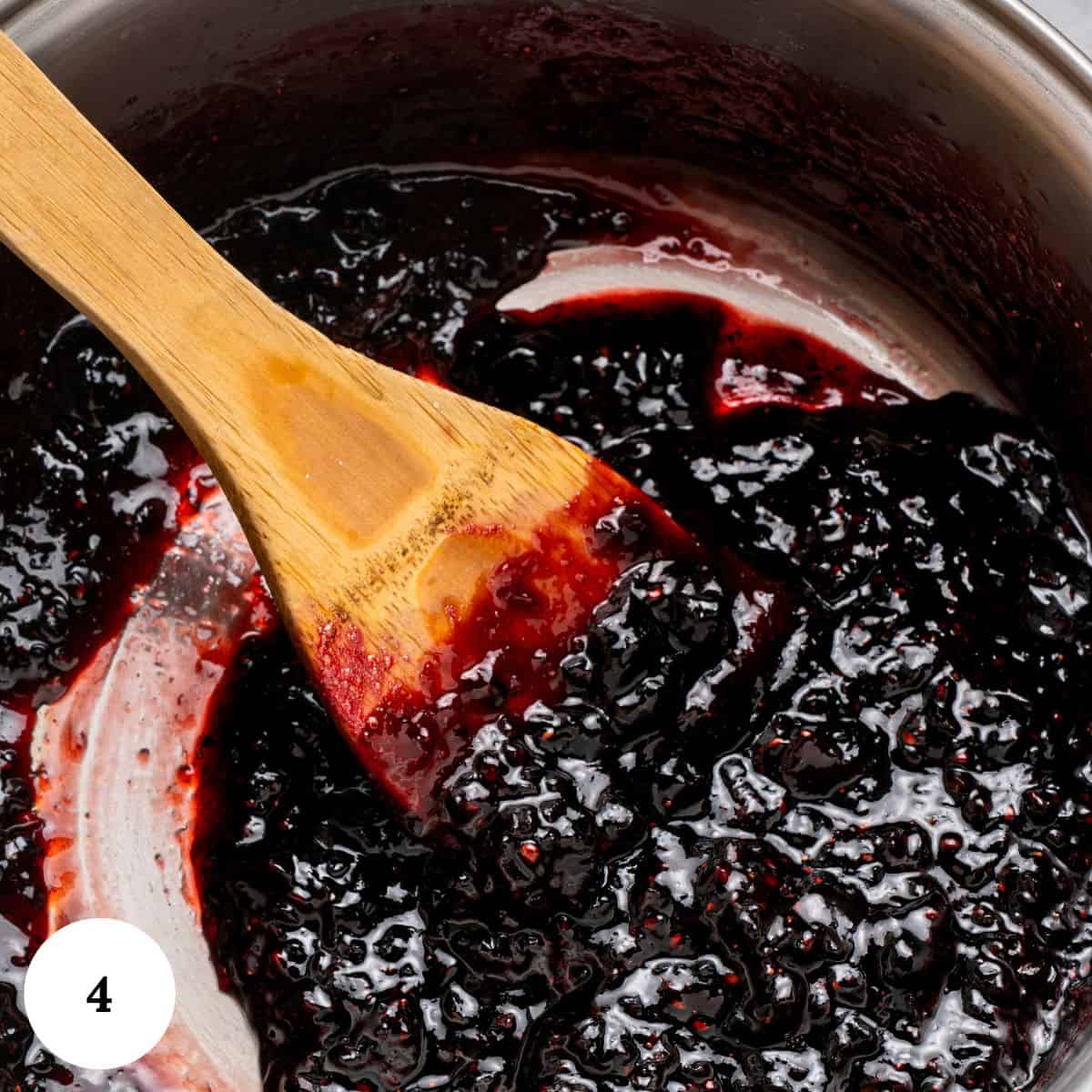 A wooden spoon stirring thick, dark purple berry pavlova jam in a metal pot. The jam has a glossy texture with visible berry pieces. A white circle with the number 4 is in the lower left corner.