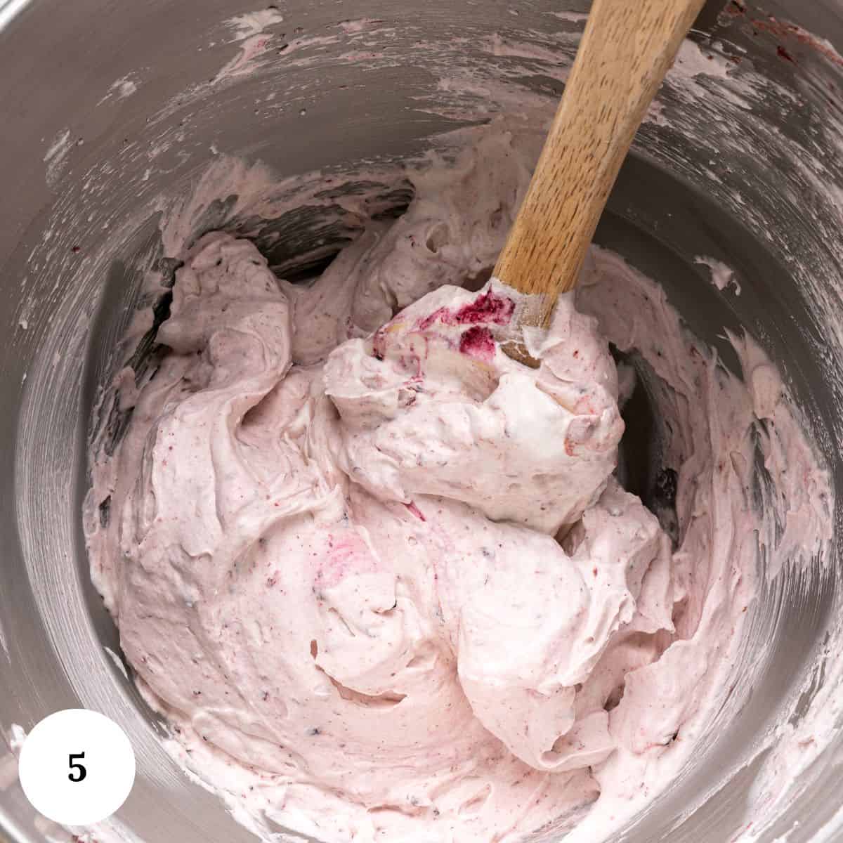 A metal mixing bowl contains a thick, pale pink creamy mixture being stirred with a wooden spatula-perfect for making berry pavlova. The mixture appears to have some berry pieces in it. A white circle with the number 5 is in the bottom left corner.