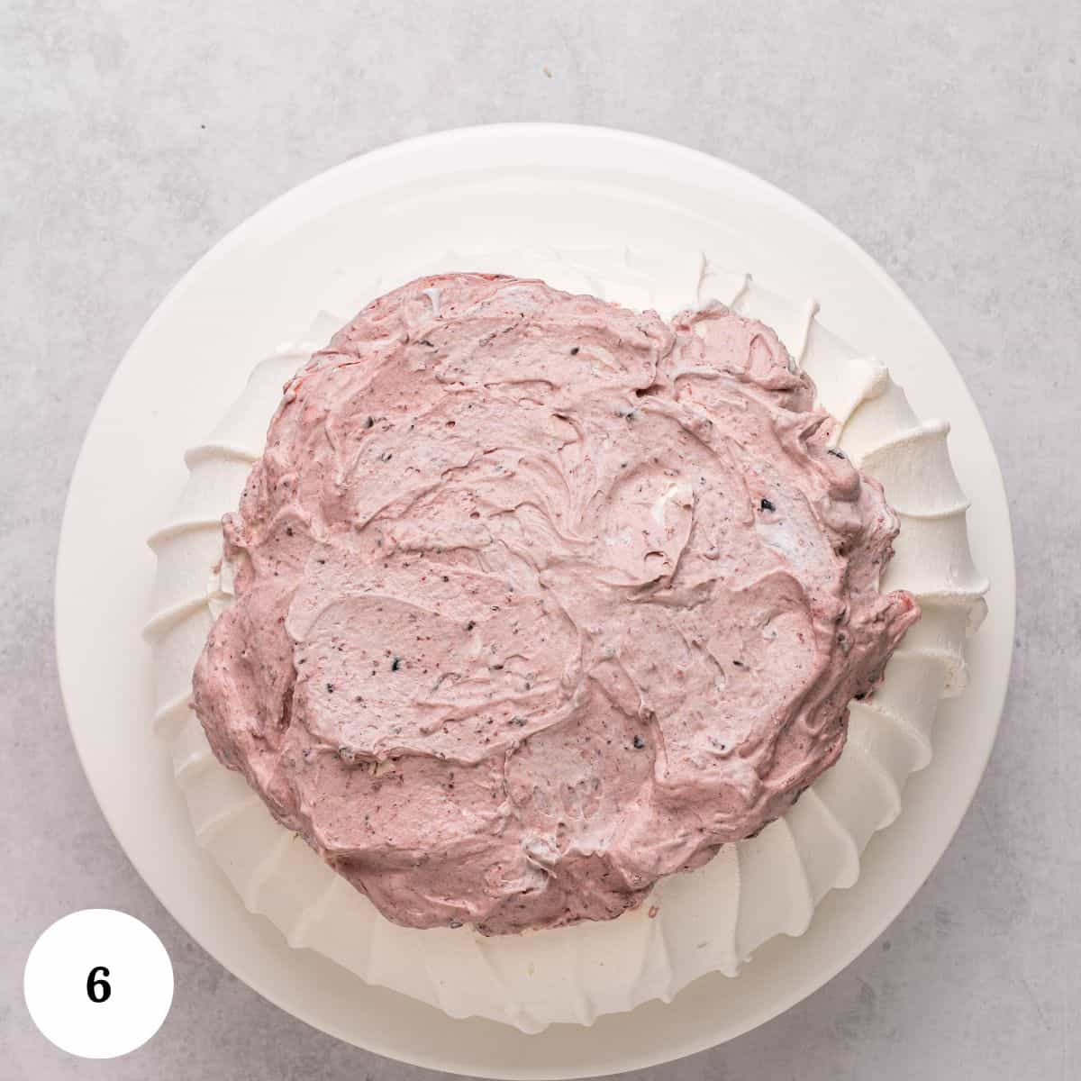 A round berry pavlova topped with a layer of light purple whipped cream sits on a white plate. The meringue base has textured, ridged edges. The image is numbered 