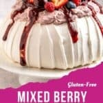 A gluten-free berry pavlova topped with whipped cream, strawberries, raspberries, blueberries, cherries, and berry sauce, displayed on a white cake stand with text overlay reading Gluten-Free Mixed Berry Pavlova.