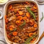 An oven roasted brisket sits in a casserole dish, topped with sliced carrots, tomatoes, and garnished with fresh rosemary and thyme. The tender meat is covered in a rich brown sauce, with plates and seasonings nearby.