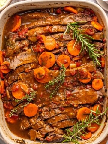An oven roasted brisket sits in a casserole dish, topped with sliced carrots, tomatoes, and garnished with fresh rosemary and thyme. The tender meat is covered in a rich brown sauce, with plates and seasonings nearby.