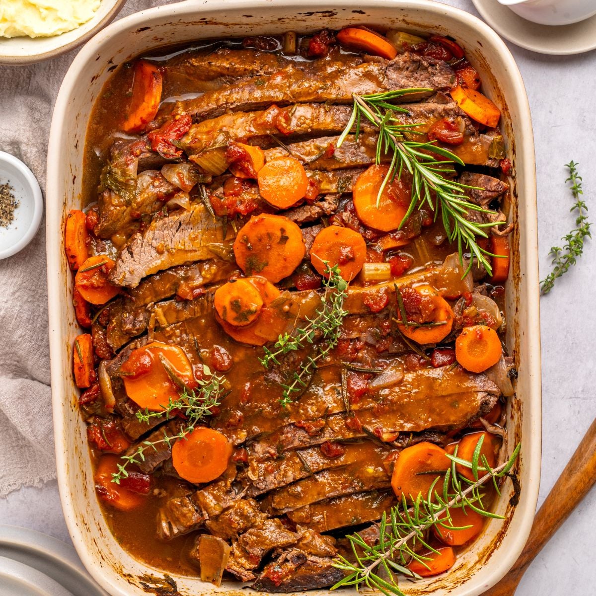 An oven roasted brisket sits in a casserole dish, topped with sliced carrots, tomatoes, and garnished with fresh rosemary and thyme. The tender meat is covered in a rich brown sauce, with plates and seasonings nearby.