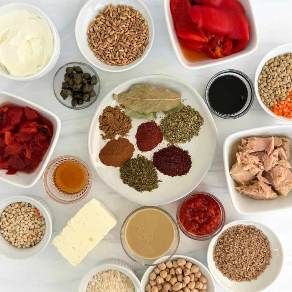 A top-down view of various small bowls containing Mediterranean pantry staples like spices, grains, legumes, vegetables, seeds, tuna, feta cheese, capers, roasted peppers, tahini, and sauces arranged neatly on a white surface.