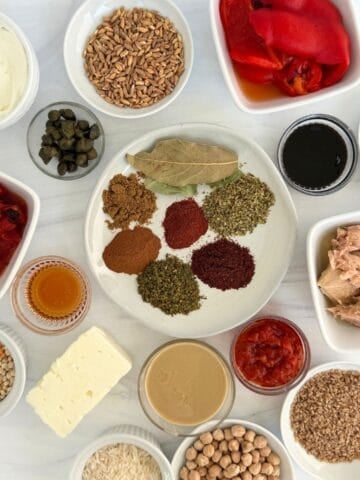 A top-down view of various small bowls containing Mediterranean pantry staples like spices, grains, legumes, vegetables, seeds, tuna, feta cheese, capers, roasted peppers, tahini, and sauces arranged neatly on a white surface.