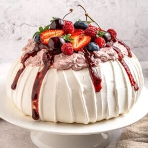 A round berry pavlova dessert with white, textured meringue topped with whipped cream, berry sauce, and fresh strawberries, cherries, blueberries, and raspberries, served on a white cake stand.