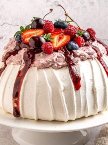 A round berry pavlova dessert with white, textured meringue topped with whipped cream, berry sauce, and fresh strawberries, cherries, blueberries, and raspberries, served on a white cake stand.