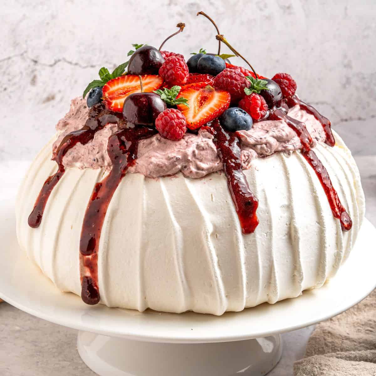 A round berry pavlova dessert with white, textured meringue topped with whipped cream, berry sauce, and fresh strawberries, cherries, blueberries, and raspberries, served on a white cake stand.