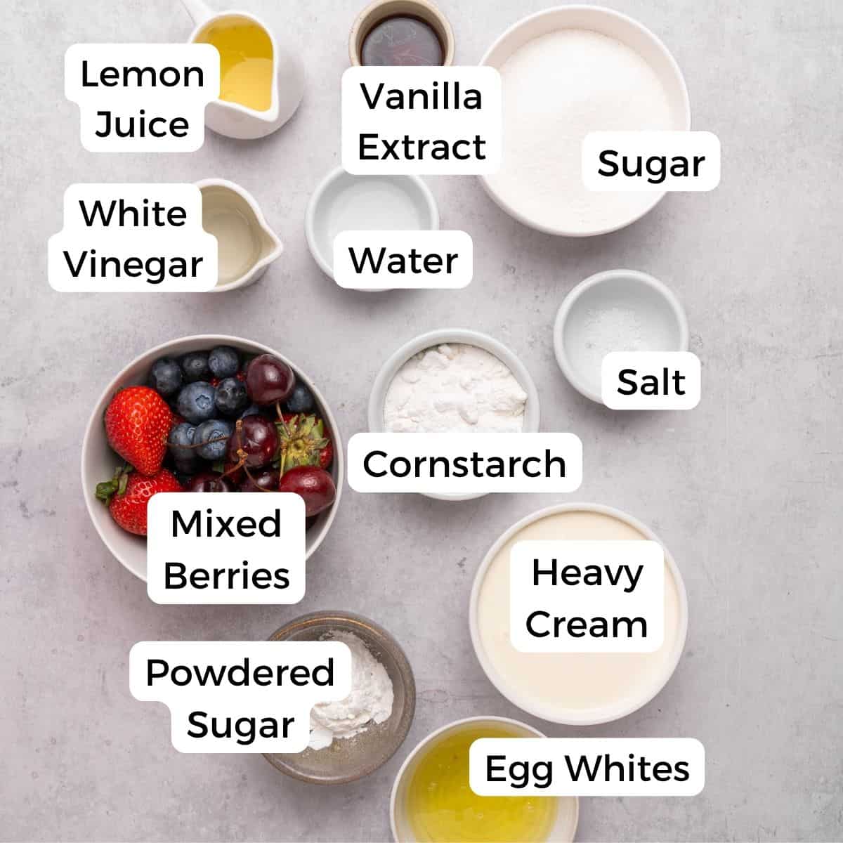 Labeled ingredients in bowls on a light surface, including those needed for berry pavlova: lemon juice, white vinegar, vanilla extract, sugar, water, salt, mixed berries, cornstarch, heavy cream, powdered sugar, and egg whites.