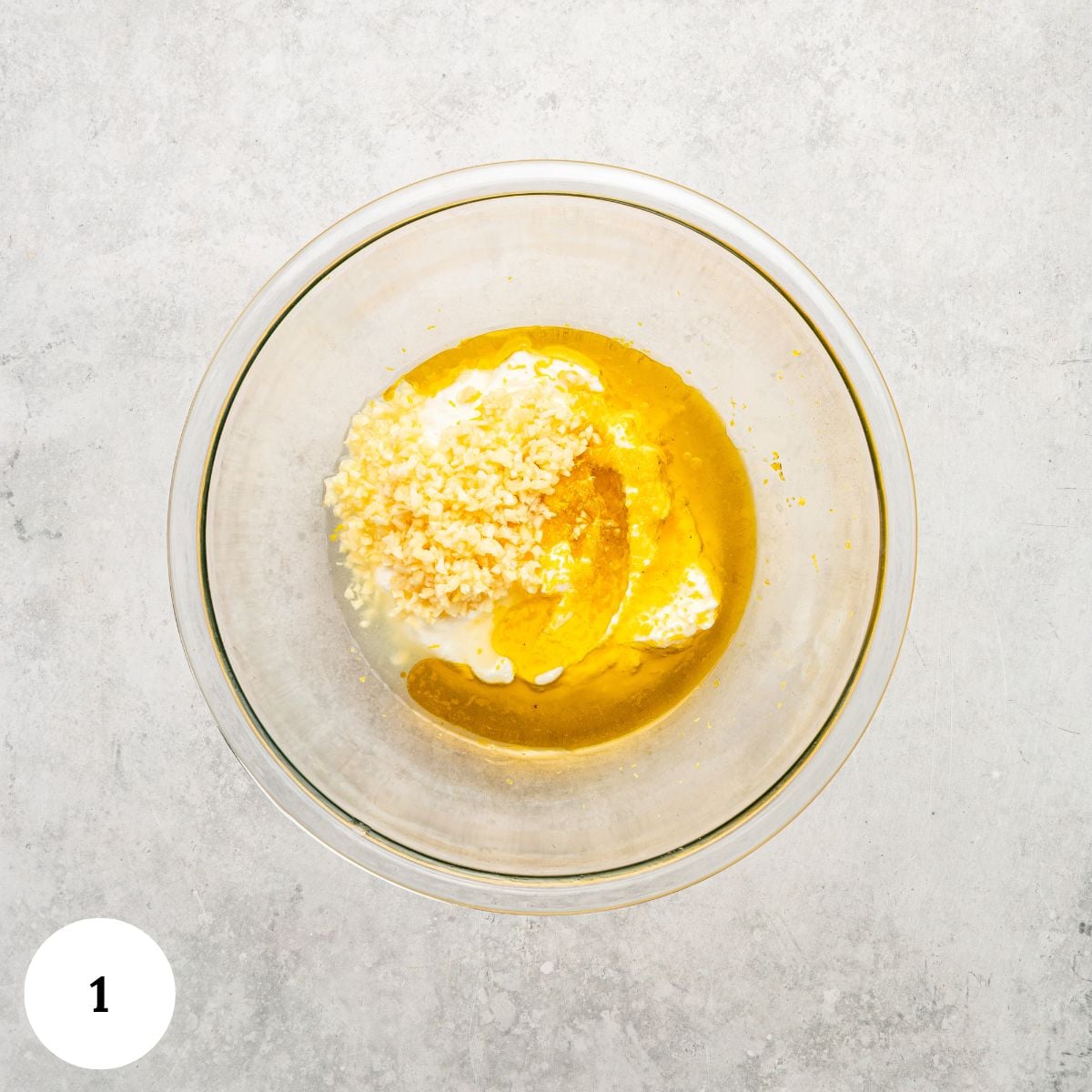 A glass mixing bowl containing yellow sauce, white cream, and minced garlic, seen from above. The bowl sits on a light gray surface-ingredients ready for Greek chicken with yogurt marinade-with a round label marked 1 in the lower left corner.