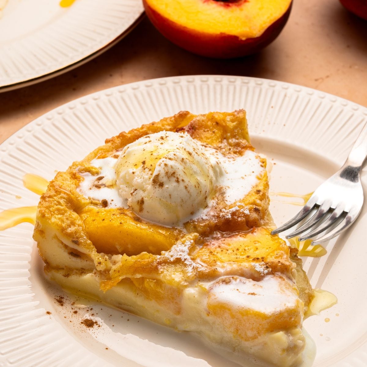 A slice of peach cobbler, reminiscent of peach clafoutis, is topped with a scoop of vanilla ice cream and a sprinkle of cinnamon, served on a white plate with a fork. Sliced peaches and another plate appear in the background.