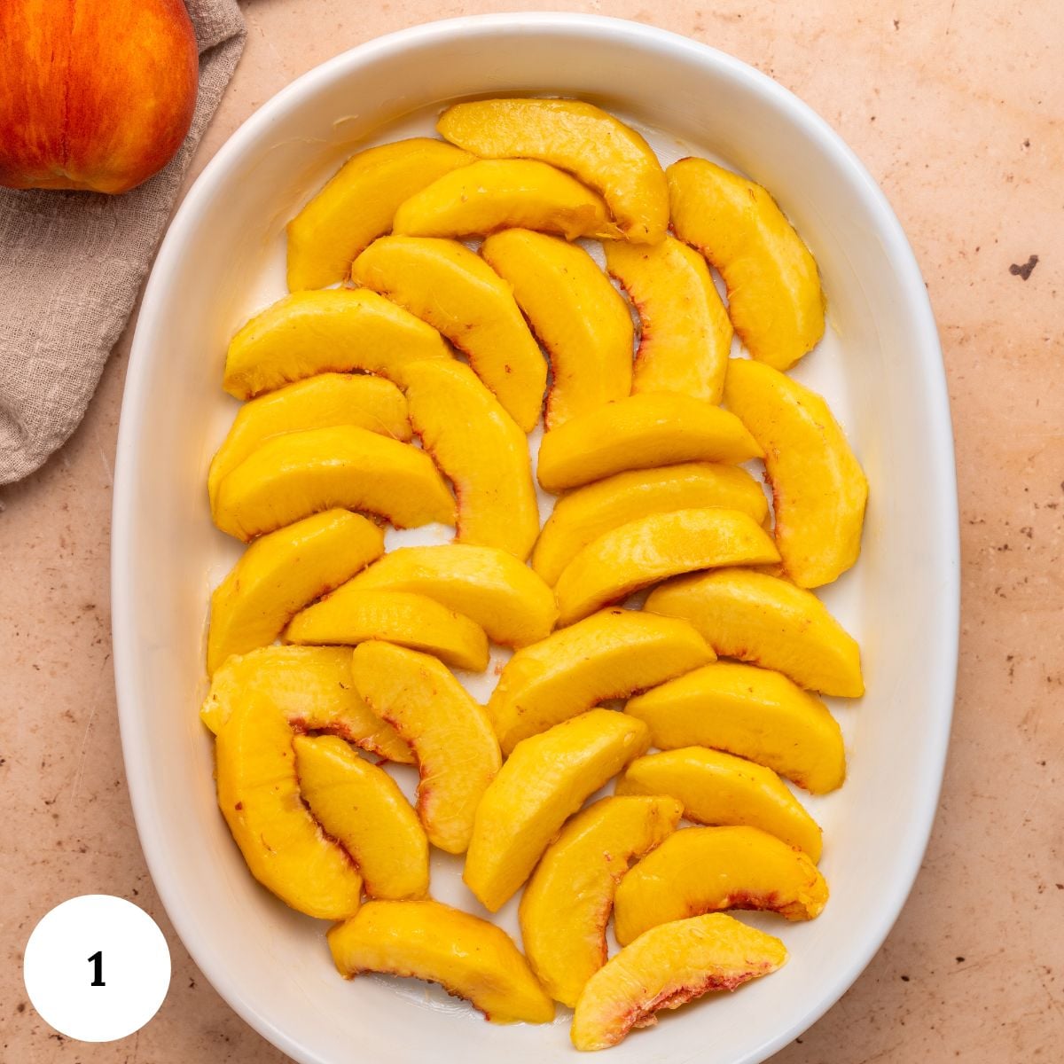 Sliced fresh peaches arranged in a single layer for peach clafoutis fill a white oval baking dish, set on a light-colored countertop with a peach and cloth napkin nearby. A white circle with the number 1 is in the lower left corner.