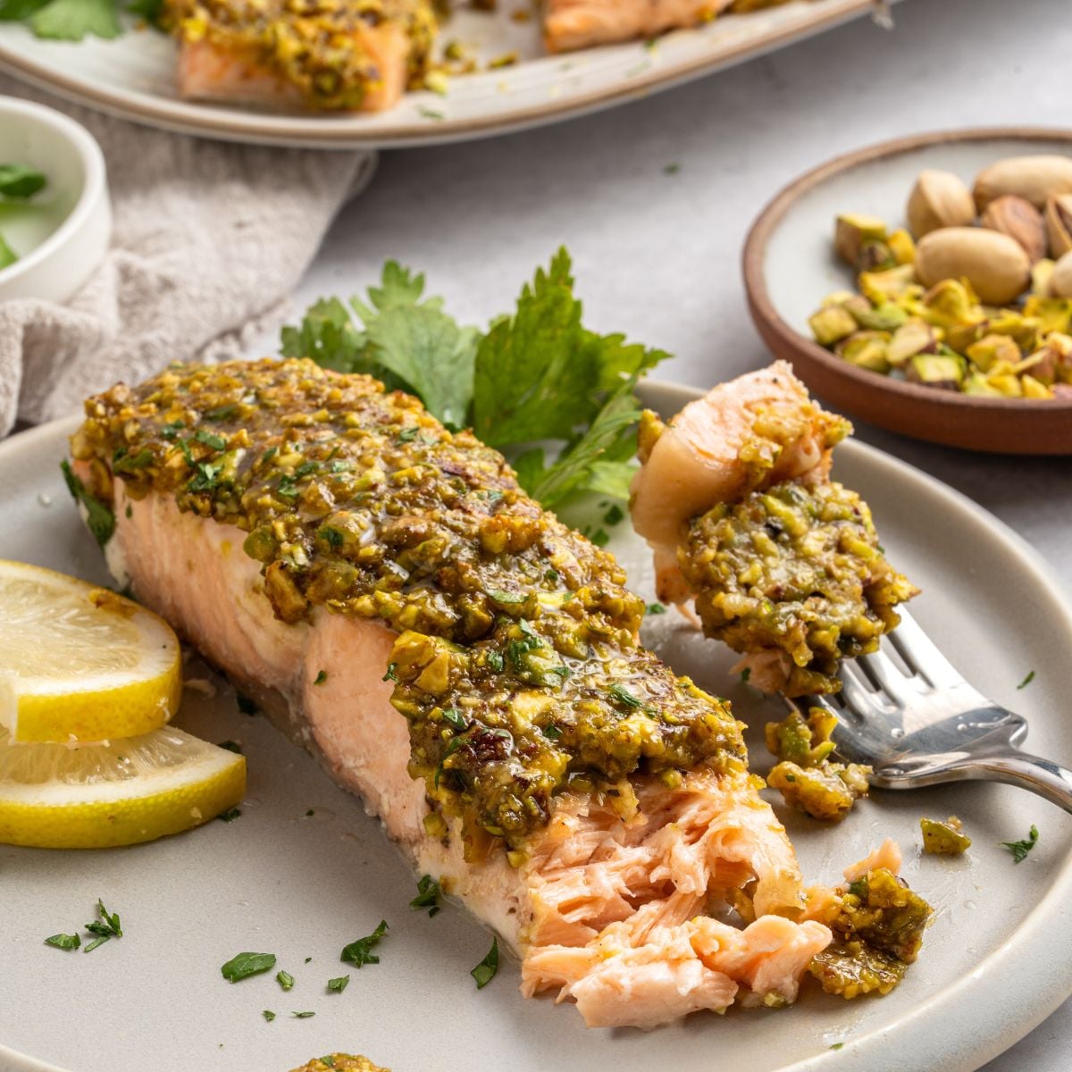A fork holds a bite of pistachio crusted salmon—a fillet topped with a chopped pistachio crust, served with lemon slices and parsley. In the background, more pistachios and another plate of fish complete the scene.