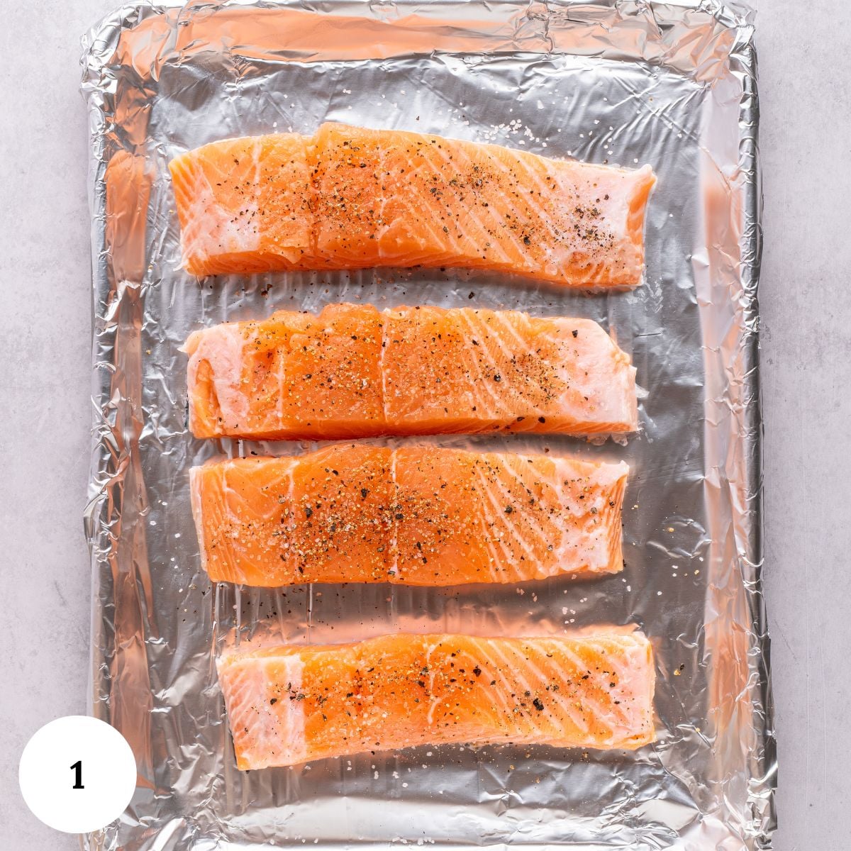 Four raw salmon fillets, ideal for making pistachio crusted salmon, are seasoned with salt and pepper and arranged in a row on a foil-lined baking sheet, ready for cooking.