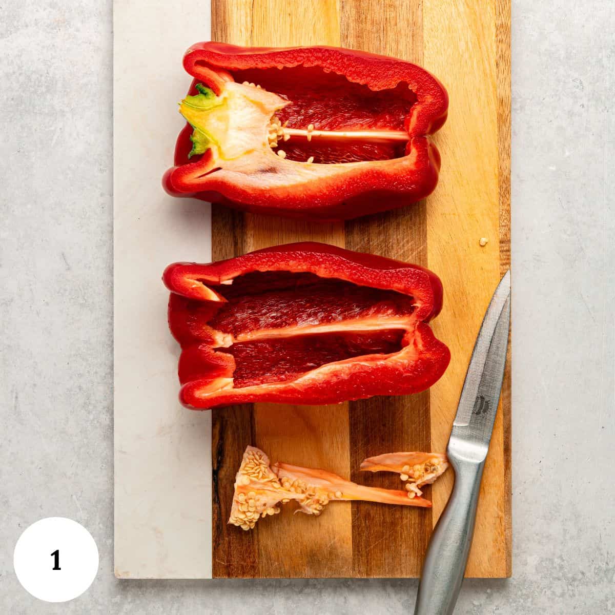 A red bell pepper sliced in half lengthwise on a wooden cutting board, with seeds and stem removed. A paring knife lies nearby-perfect for prepping Roasted Red Bell Pepper Hummus. Light background, white number 1 at the bottom left.