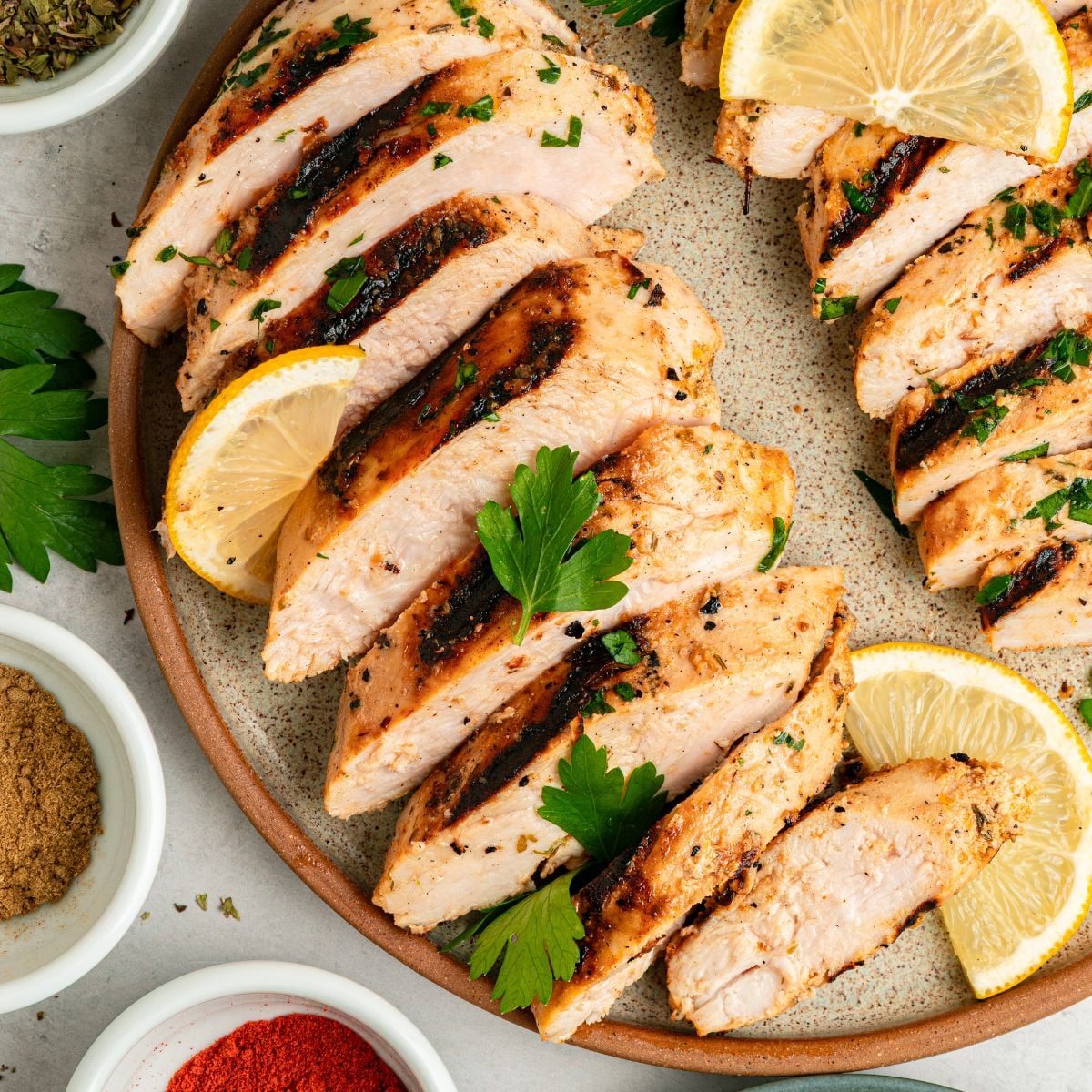 Sliced grilled Greek chicken with yogurt marinade, garnished with fresh parsley and lemon wedges, served on a plate with small bowls of ground spices nearby.