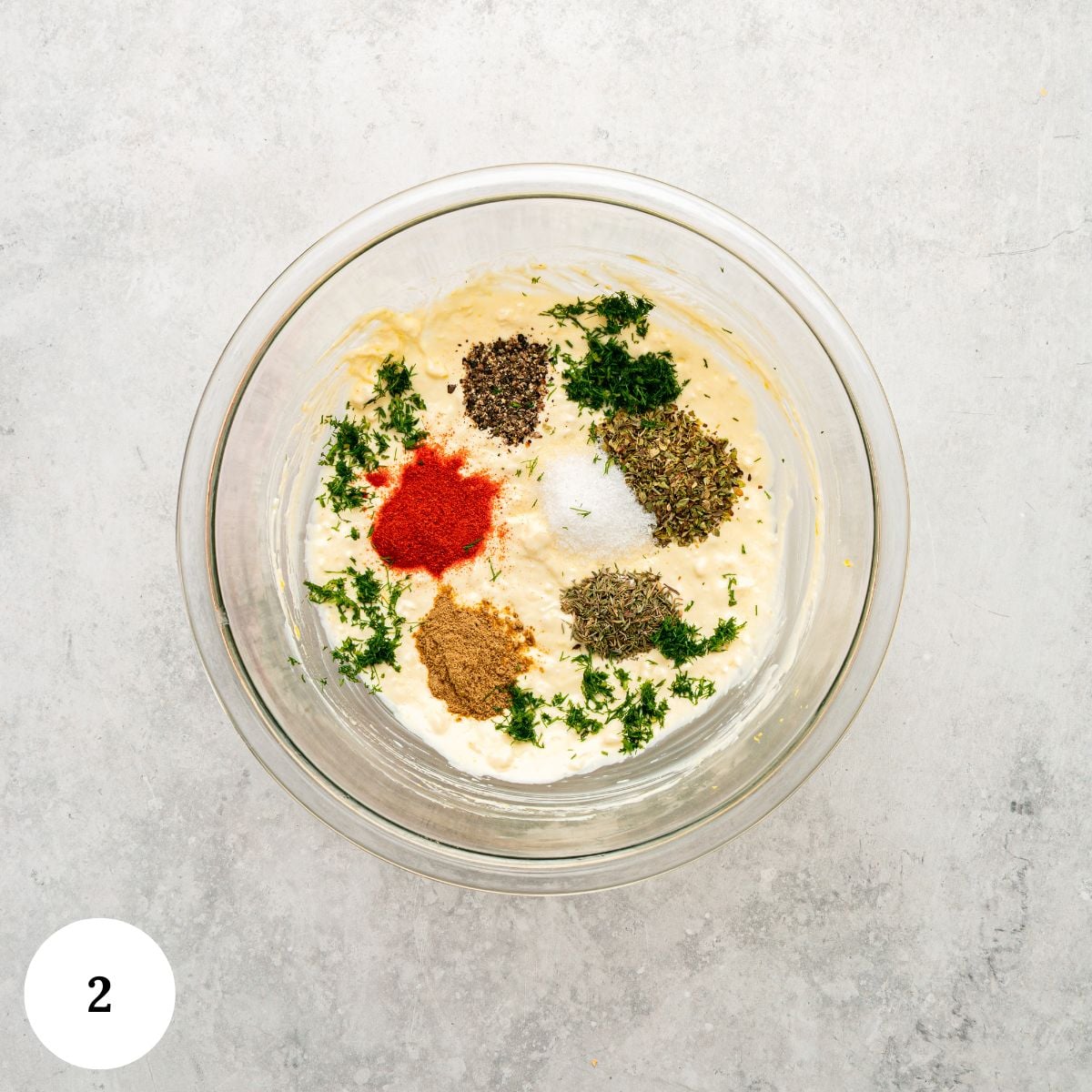 A glass bowl with creamy greek chicken with yogurt marinade, topped with piles of black pepper, dried herbs, red paprika, green herbs, and ground spices sits on a light gray surface. A white circle with the number 2 is in the bottom left corner.