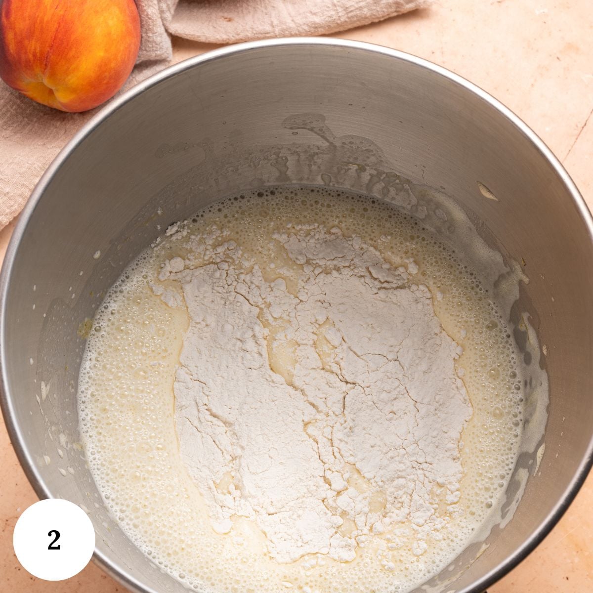 A mixing bowl containing wet batter with flour on top, ready to be mixed for a peach clafoutis. A peach and a beige towel are visible nearby on a light countertop. A white circle with the number 2 is in the lower left corner.
