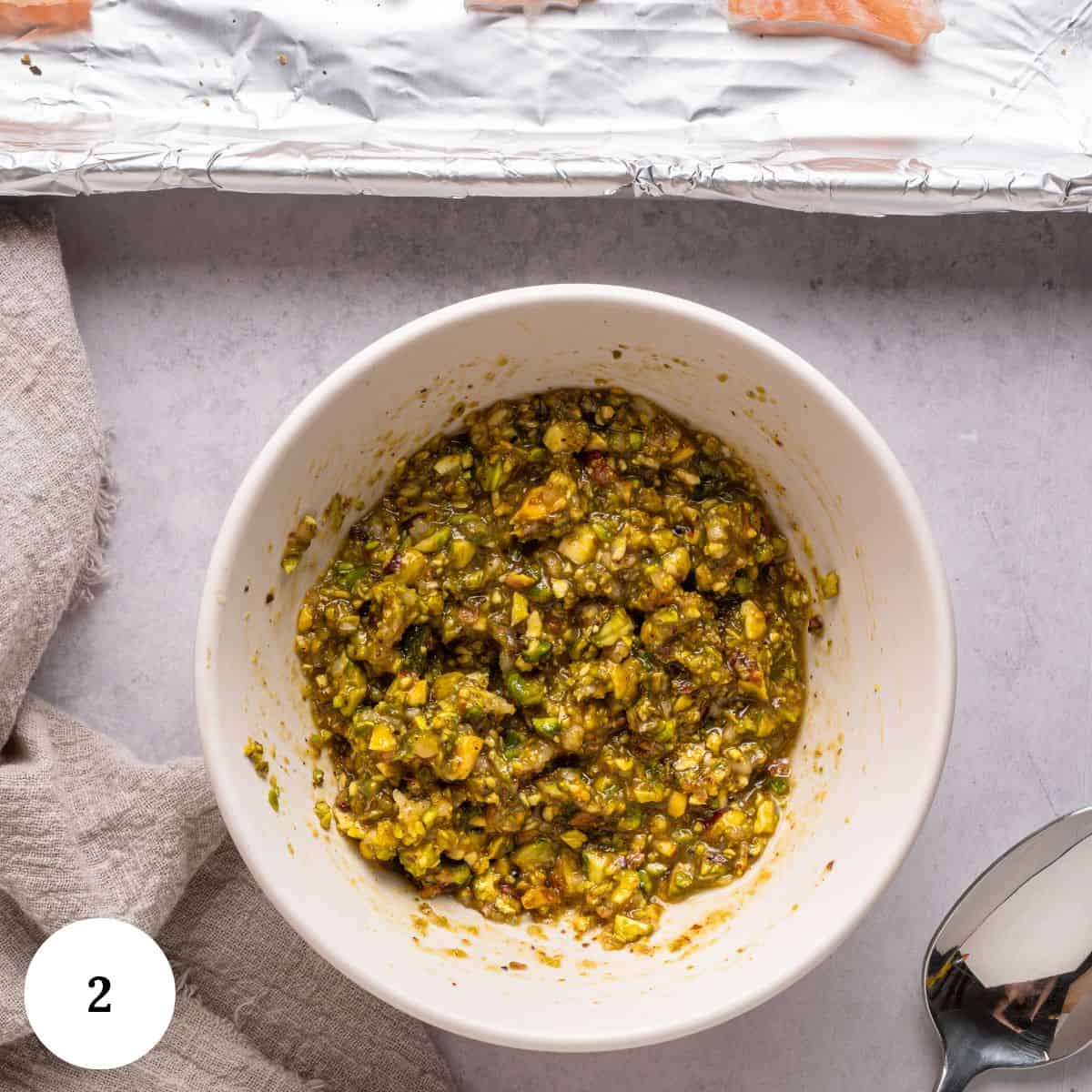 A white bowl filled with a chunky green and yellow pesto mixture sits on a light countertop next to a silver spoon, a beige cloth, and a foil-lined baking sheet with pistachio crusted salmon pieces. A 2 label is in the bottom left corner.