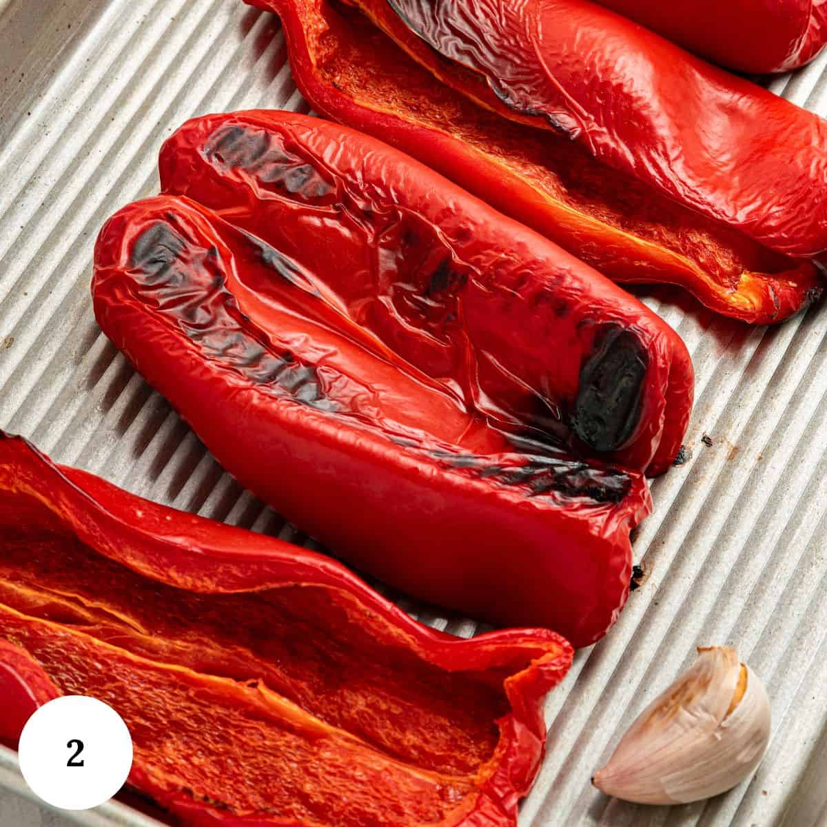 Roasted Red Bell Pepper Hummus features charred red bell pepper halves and garlic on a metal tray. A white circle with the number 2 is in the lower left corner.