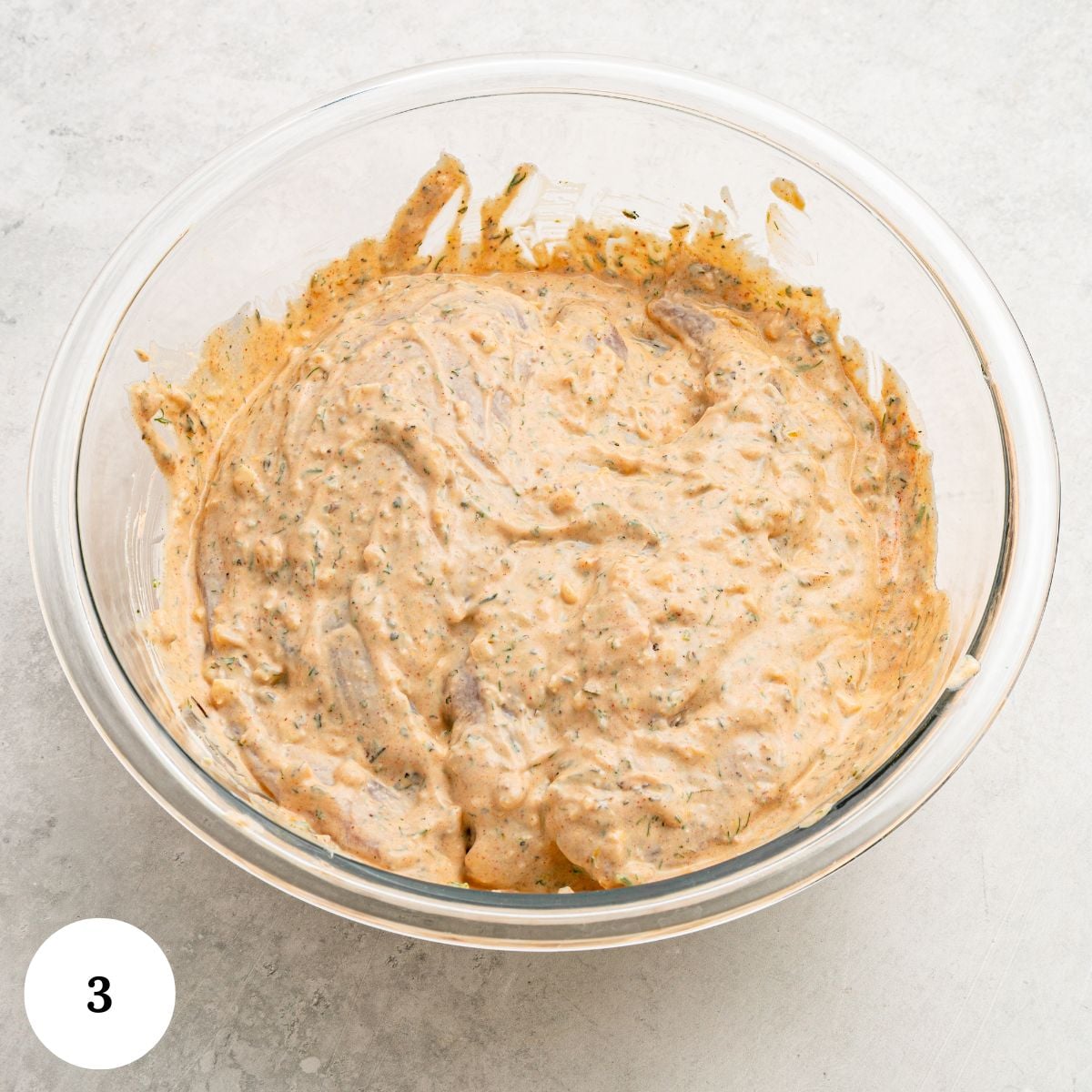 A glass bowl filled with a creamy, orange-colored Greek chicken with yogurt marinade, flecked with herbs and spices, sits on a light gray surface. A white circle with the number 3 appears in the bottom left corner.