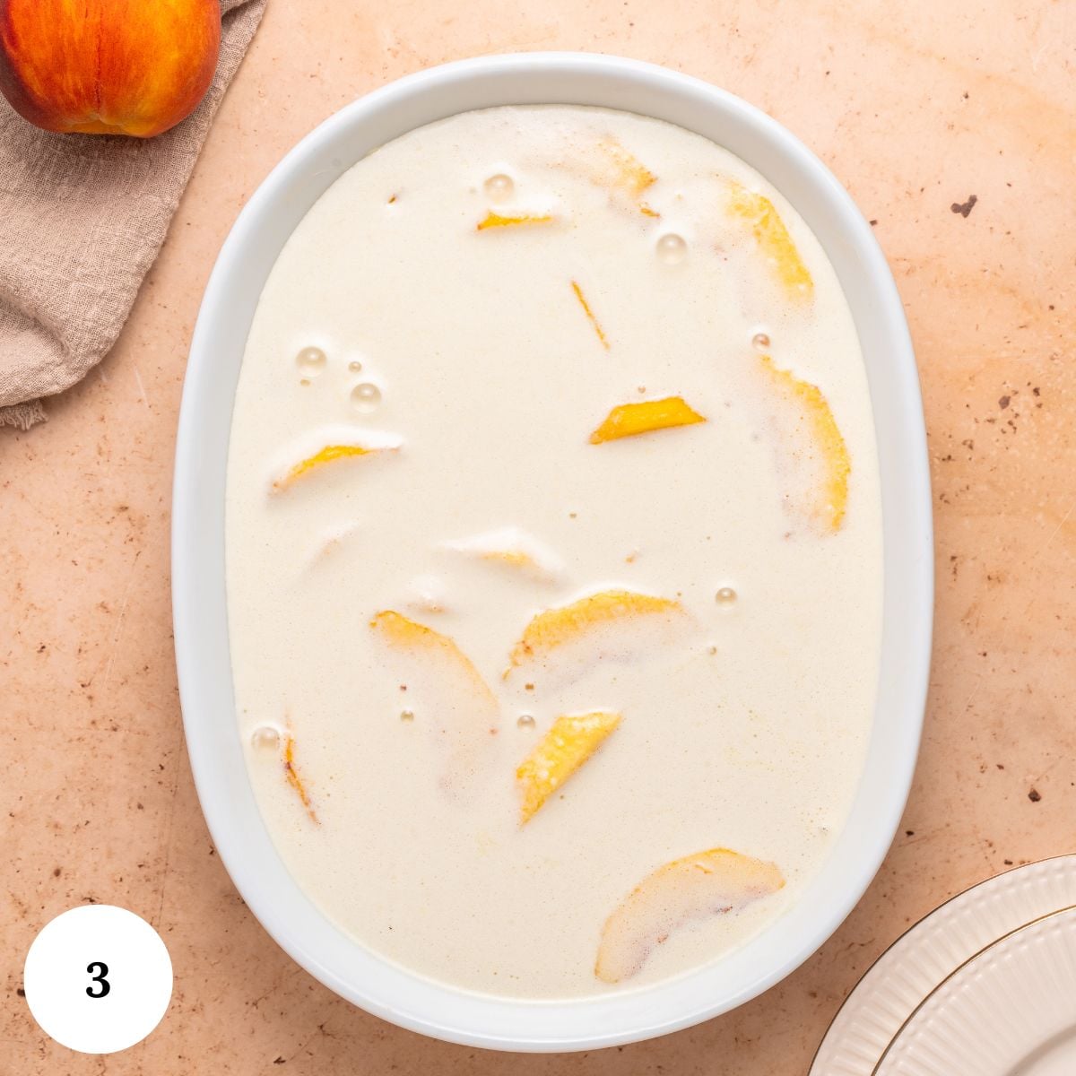 A white oval baking dish filled with sliced peaches and creamy batter-perfect for a peach clafoutis-sits on a light surface. A fresh peach and beige cloth are nearby, with the number 3 in a white circle in the corner.