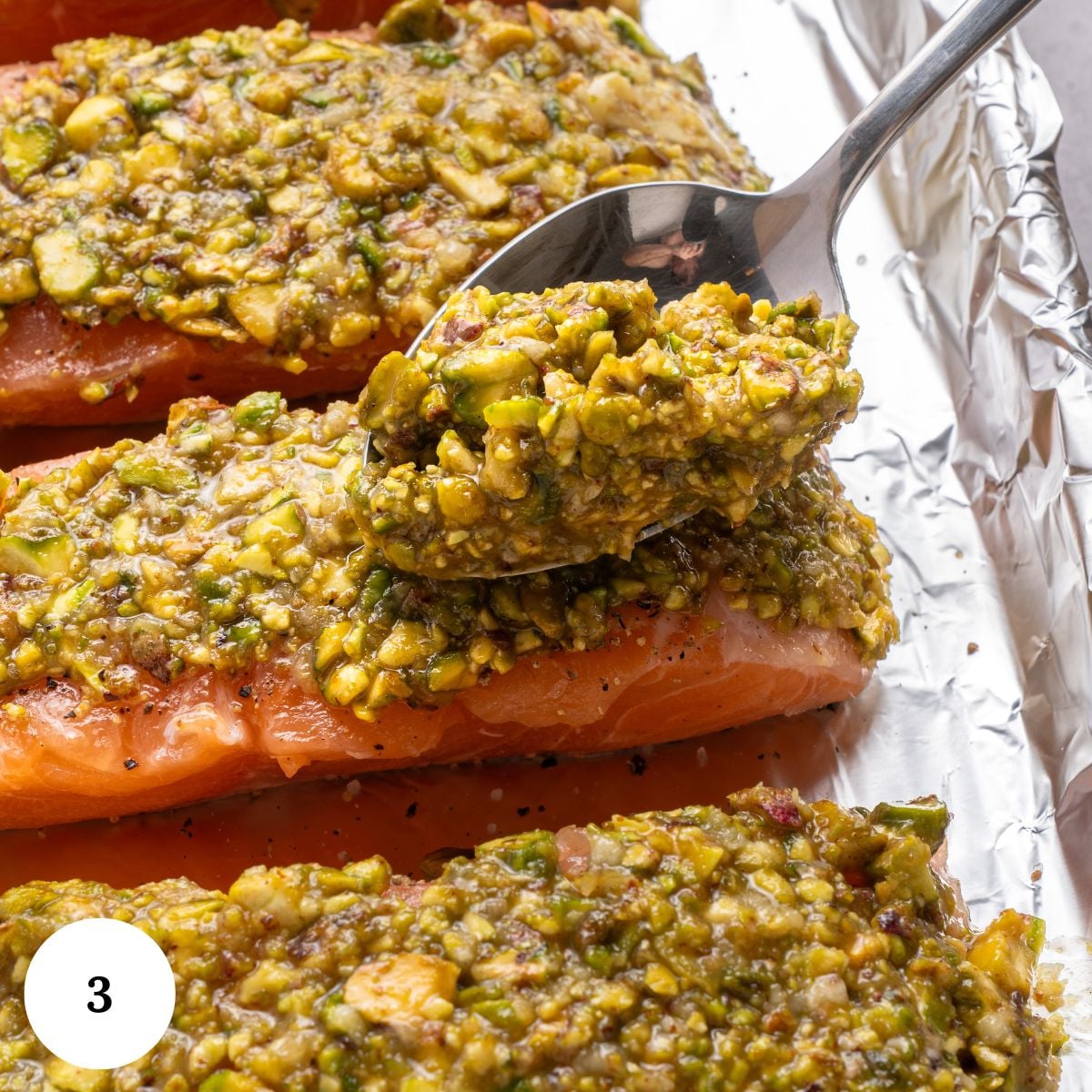 A spoon spreads a chunky, greenish pistachio crust over raw salmon fillets, creating delicious pistachio crusted salmon on a foil-lined baking tray. A white circle with the number 3 appears in the corner.