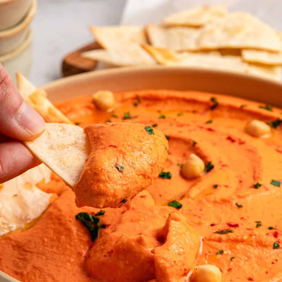 A hand dipping a pita chip into a bowl of creamy Roasted Red Bell Pepper Hummus, garnished with chickpeas, chopped herbs, and paprika. More pita chips are visible in the background.