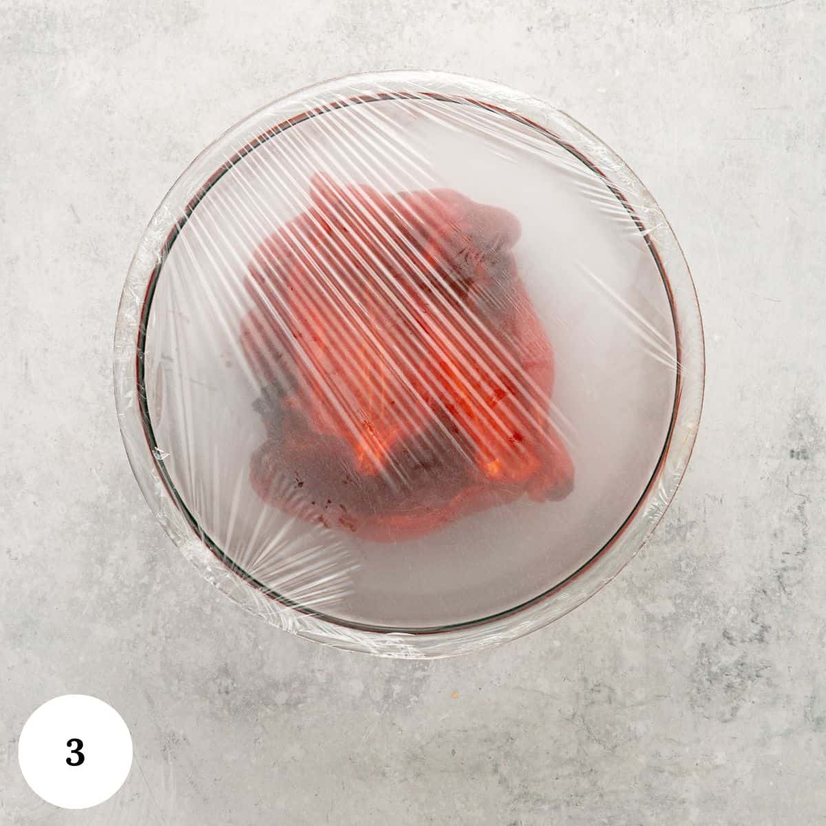A glass bowl covered with plastic wrap, containing a red roasted pepper for Roasted Red Bell Pepper Hummus, sits on a light gray surface. The number 3 is displayed in a white circle in the bottom left corner.