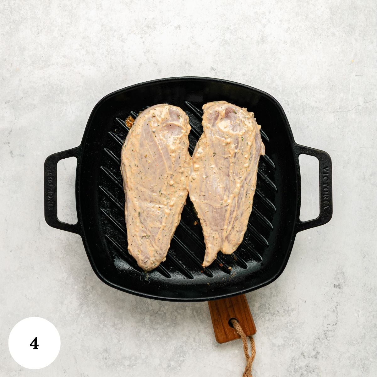 Two raw greek chicken breasts with yogurt marinade are placed side by side in a black grill pan with handles on a light gray surface. A wooden holder is attached to the pan, and a white circle with the number 4 is in the bottom left corner.