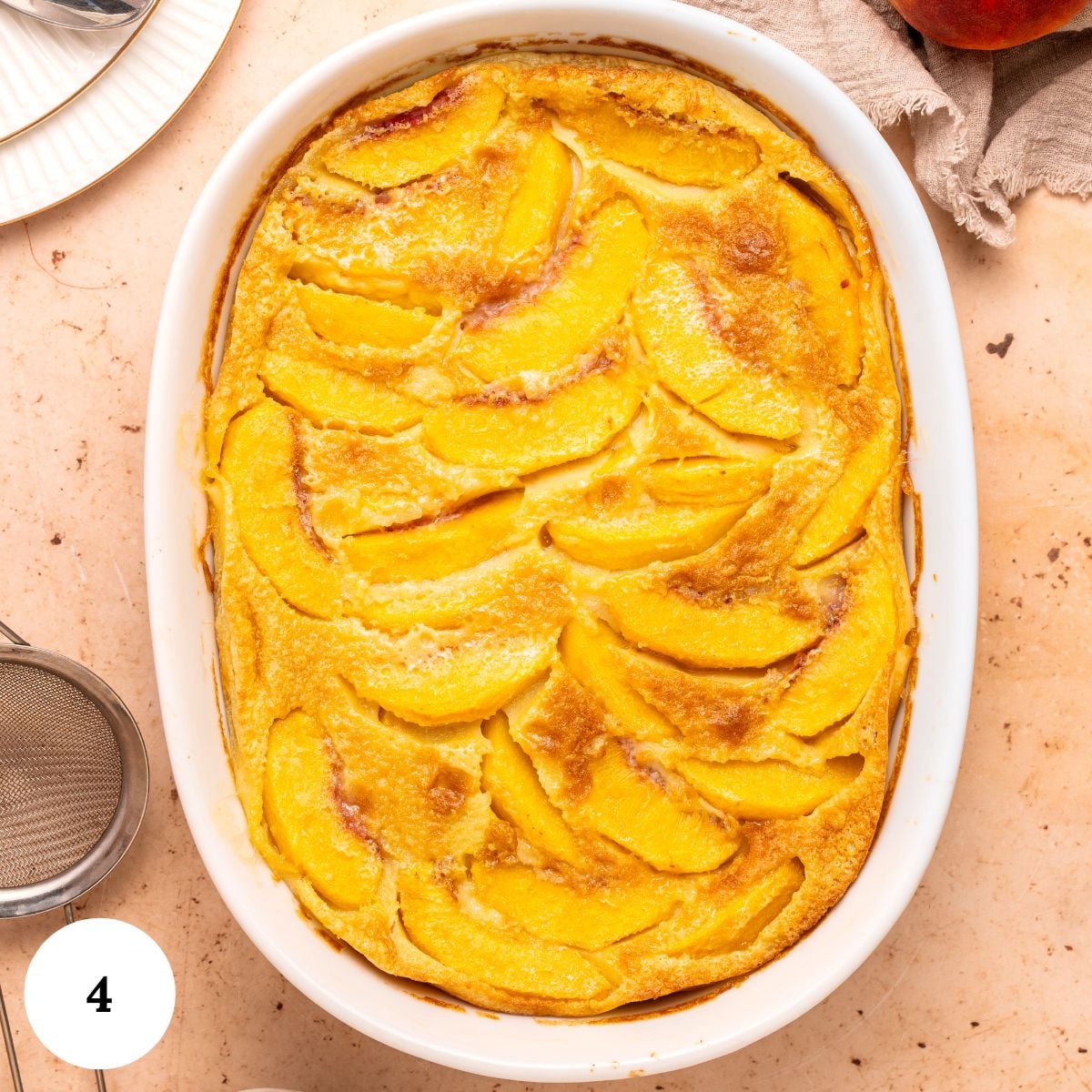 Baked peach clafoutis in a white oval dish, topped with golden-brown peach slices, sits on a light countertop next to a sifter and a plate.