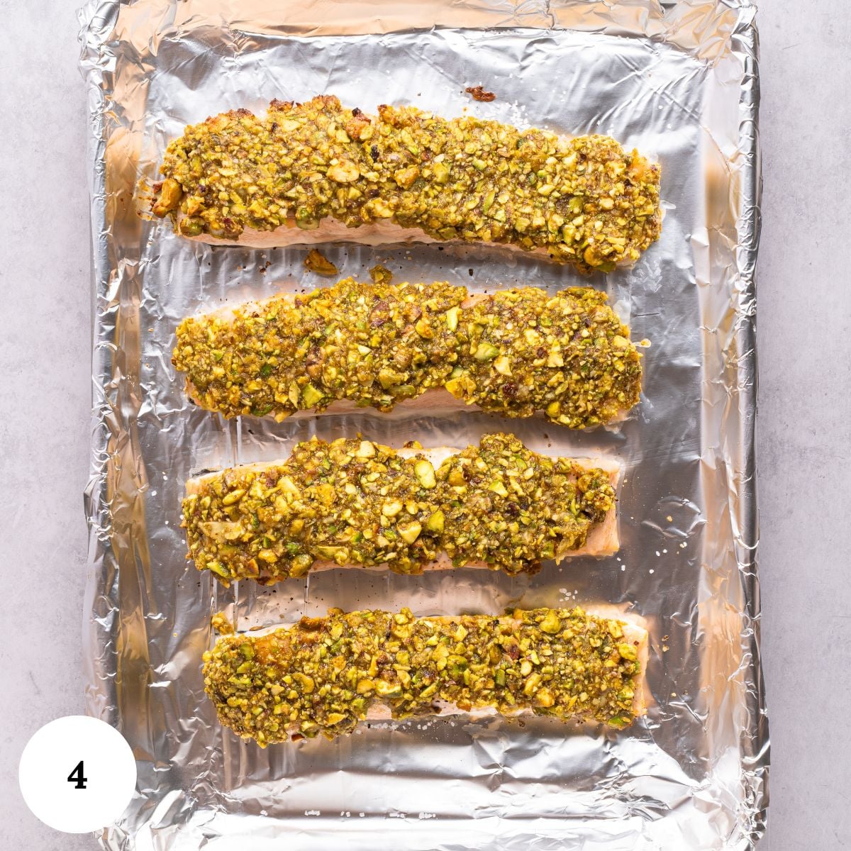 Four pistachio crusted salmon fillets are coated in a crushed pistachio crust and arranged on a foil-lined baking sheet. The image is shot from above on a light background, with a small white circle labeled 4 in the bottom left corner.